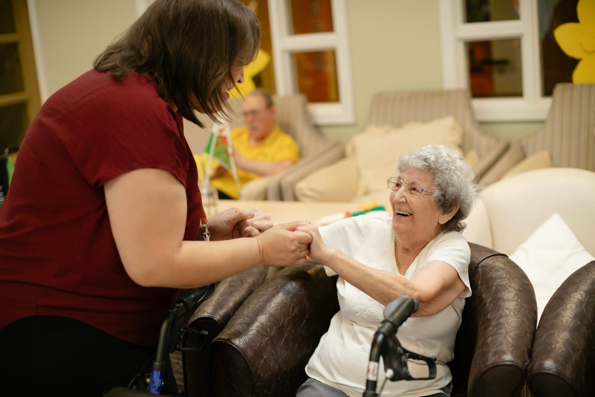 A caregiver holding an older woman's hands in her living room.